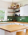 ELMHURST, IL, USA - MAY 25, 2021: A renovated kitchen with white cabinets, a natural wood countertop, chairs sitting at the island, a green subway tile backsplash, and a gold pendant light.