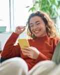 Happy relaxed young woman sitting on couch using cell phone, smiling lady laughing holding smartphone, looking at cellphone enjoying doing online ecommerce shopping in mobile apps or watching videos.