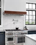 A luxurious kitchen with blue cabinets, a stainless steel stove, granite backsplash and countertop, and black framed windows.