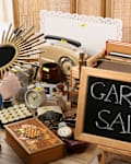 Woman with beautiful mirror at table. Sign Garage Sale written on blackboard near many different stuff indoors