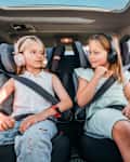 Portrait of two positive smiling sisters sitting in child car seats fastening safety belts listening to music using headphones. Happy childhood, auto journey concept image.