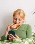 young woman lying in bed with a phone and munching on toast