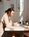 Minimal side view portrait of young woman writing in notebook at home with sun streaming in.