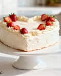 A beautiful heart-shaped cake, frosted with white cream and topped with fresh strawberries, sits on a stand in a modern, sunlit kitchen.