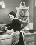 Woman in vintage kitchen washing dishes, wearing a floral apron, with shelves displaying teapot and plates.