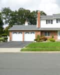 Two-story suburban house with brick and siding exterior, two-car garage, manicured lawn, and shrubs.