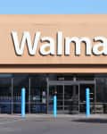 Walmart storefront with large logo, glass entrance, blue bollards, and a tree on the right.