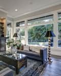 living room filled with built-in cabinets flanking round mirror atop grey tile fireplace, tufted sofa facing two armchairs and window wall overlooking lush outdoors
