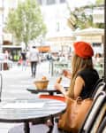 Young stylish woman in red beret having a french breakfast with coffee and croissant sitting oudoors at the cafe terrace