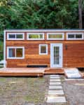 Tiny house with wood siding, white-framed windows, and a small deck with two chairs, set in a forested area.