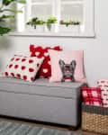 Gray bench with heart-patterned pillows, a dog print pillow, potted plant, and woven basket in a cozy room.
