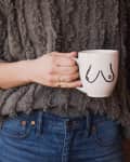 Person holding a white mug with a minimalist breast illustration, wearing a textured gray sweater and blue jeans.