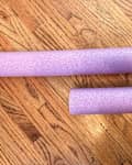 Two purple foam pool noodles resting on a wooden floor, one longer and one shorter.