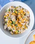 Bowl of chicken salad with corn, peas, and crushed chips, next to a plate of crispy snacks on a light surface.