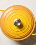 Round yellow cast iron Dutch oven with a stainless steel knob on a marble surface.