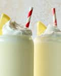 Two creamy lemon drinks topped with whipped cream and lemon slices, each with a red and white striped straw.