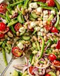 A colorful salad with cherry tomatoes, green bell peppers, cucumbers, red onion, feta cheese, and white beans, garnished with herbs.