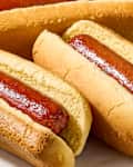 Three hot dogs in soft buns on a white plate, showcasing shiny sausage and golden bread.