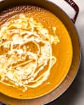 Image 2: Creamy orange soup in a deep pot, swirled with cream and topped with black pepper.