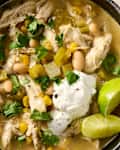 Bowl of chicken soup with shredded chicken, white beans, corn, green chilies, cilantro, and a dollop of sour cream, garnished with lime.