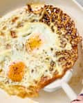 Two fried eggs in a cream-colored skillet, one with crispy edges, seasoned with black pepper.