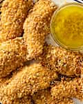 Crispy breaded chicken tenders arranged on a plate with a small bowl of yellow dipping sauce.