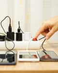 Hand turning on power strip with multiple chargers and devices on a wooden table.