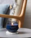 Blue candle labeled "Warm Luxe Cashmere" on a round gray table, with a wooden chair and blue pillow in the background.