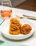 Spaghetti and meatball in white bowl on marble table with a glass of rosé wine and gold cutlery.