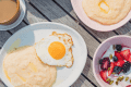 Breakfast spread with fried eggs, grits, yogurt with berries, coffee, and pastries on a wooden table.