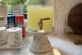 Glass beverage dispenser with yellow liquid on a marble stand, next to spice jars and canned drinks on a kitchen counter.