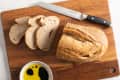 Sliced bread on a wooden cutting board with a serrated knife and a bowl of olive oil and balsamic vinegar.