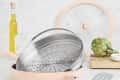 Pink nonstick pan with steamer insert, spatula, olive oil, artichoke, and salt on a kitchen counter.