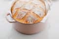 Freshly baked bread in a pink Dutch oven on a marble countertop, with butter and herbs on a tray in the background.