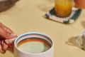 Hand holding a white mug with colorful rim on a yellow tablecloth, next to a glass of orange juice.