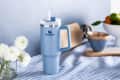 Blue Stanley mug with handle and straw on striped cloth, next to white flowers and kitchen items.