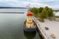 Tugboat with a smiling face and red cap docked at a waterfront, surrounded by trees and sailboats in the background.