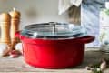 Red Dutch oven with glass lid on wooden table, surrounded by garlic, pepper mills, and olive oil can.