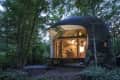 Curved wooden cabin with large windows, nestled in a forest, interior warmly lit.
