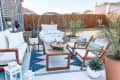 Outdoor patio with wooden furniture, blue and white rug, potted plants, and decorative lanterns.