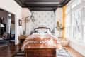 Bedroom with patterned wallpaper, wooden chest, orange throw, large windows, and yellow curtains.
