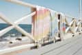 Pastel-striped towels draped over a white wooden railing by the beach with ocean waves in the background.