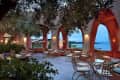 Outdoor terrace with arched openings, metal chairs, and tables, overlooking a scenic view of the sea and distant mountains.