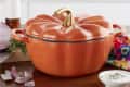 Orange pumpkin-shaped casserole dish with a gold stem handle on a wooden countertop, surrounded by sliced onions and herbs.
