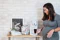 Pregnant woman in gray dress using a coffee maker on a wooden shelf with mugs, jars, and decor against a brick wall.