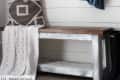 Round mirror and hooks on shiplap wall, denim shirt hanging, bench with patterned pillow and white throw.