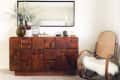 Wooden sideboard with geometric design, large mirror, vase with foliage, and a cane rocking chair with a white fur cushion.