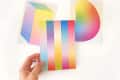Hand holding a colorful gradient postcard with two abstract gradient art prints in the background.