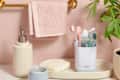 bathroom sink scene with a soap dispenser, lit candle, bar soap, and a white toothbrush organizer with toothbrushes and toothpaste in them against a peach wall with a plant on the right