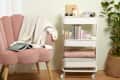 living room vignette with a white rolling cart being used to organize blankets, books, and art supplies between a chair with a blanket draped over it and a plant